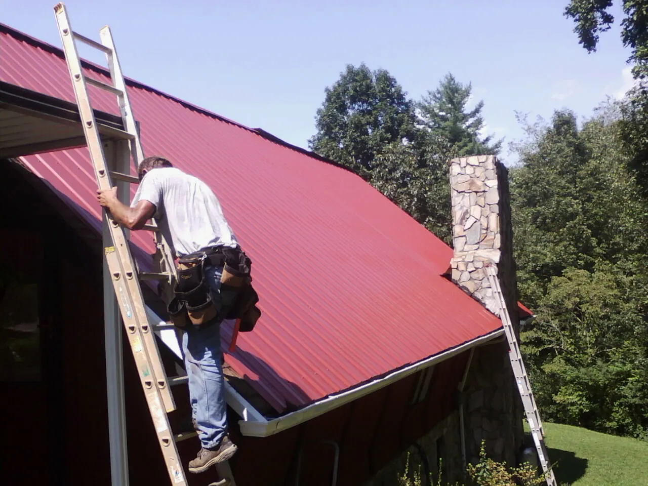 Roofer on a ladder beside a red metal roof and stone chimney — Soffit & Fascia Repair by Roofing Brothers in Bankston